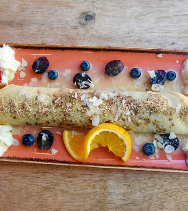 Das Bild zeigt einen Teller mit einer gefüllten Palatschinke, die auf einem farbigen, rechteckigen Teller präsentiert ist. Die Palatschinke ist rötlich-braun und aufgerollt, mit einer Mischung aus gehackten, gerösteten Nüssen und mandelartigen Streuseln bestreut. Um die Palatschinke herum befinden sich mehrere frische Früchte: zwei Erdbeeren, einige Heidelbeeren und eine halbe orange Scheibe. Auf einer Seite des Tellers gibt es einen Klecks Sahne, der in zwei Portionen verteilt ist. Die Unterlage des Tellers zeigt eine Holzoberfläche, die zur rustikalen Präsentation beiträgt.