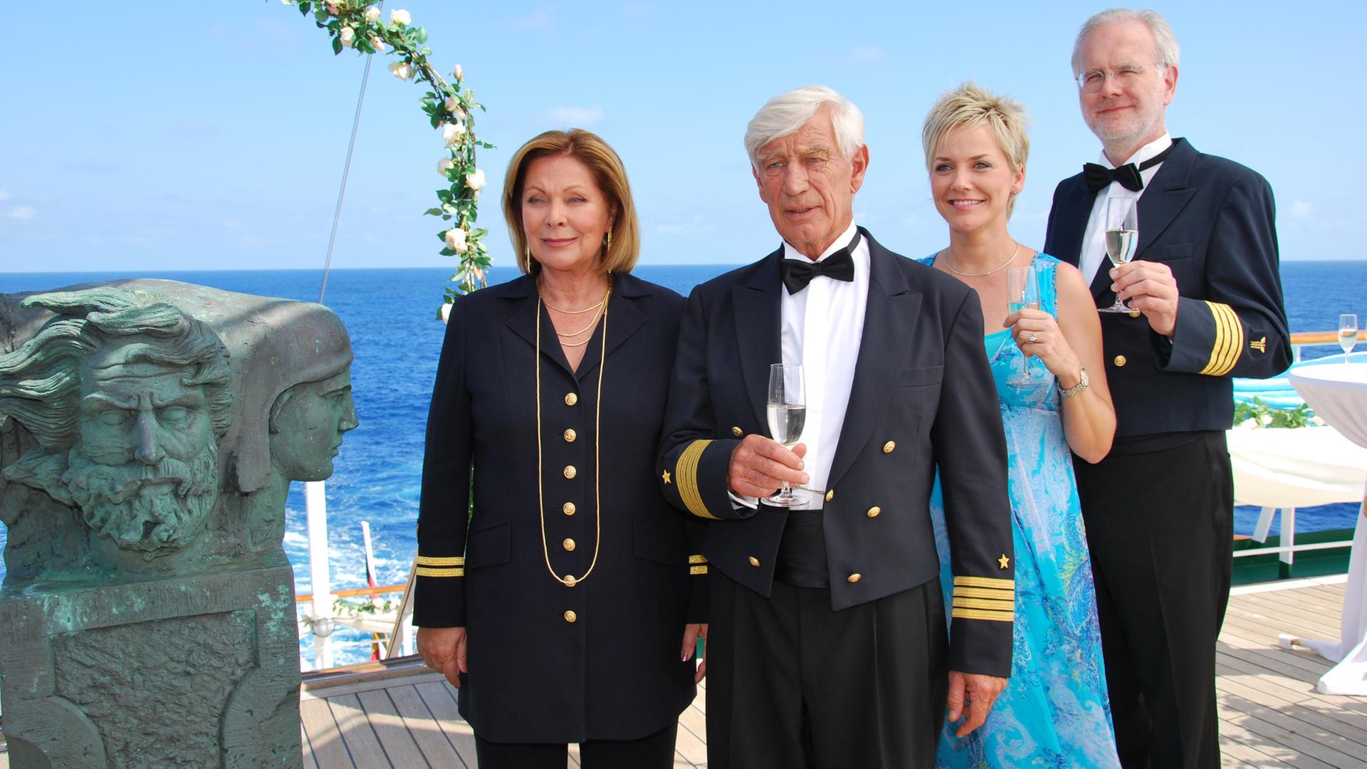 "Das Traumschiff - Panama": In feierlicher Garderobe stehen Kapitän Paulsen (Siegfried Rauch), Beatrice (Heide Keller), Inka (Inka Bause) und Oskar Schifferle (Harald Schmidt) an Deck des Traumschiffs, das mit Blumen geschmückt ist. Bis auf Beatrice halten alle ein Sektglas in der Hand und blicken in eine Richtung.