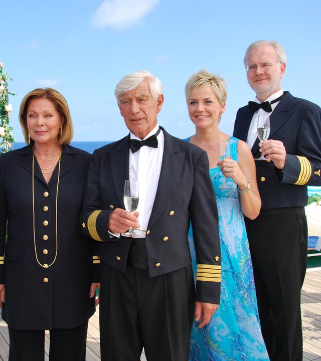 "Das Traumschiff - Panama": In feierlicher Garderobe stehen Kapitän Paulsen (Siegfried Rauch), Beatrice (Heide Keller), Inka (Inka Bause) und Oskar Schifferle (Harald Schmidt) an Deck des Traumschiffs, das mit Blumen geschmückt ist. Bis auf Beatrice halten alle ein Sektglas in der Hand und blicken in eine Richtung.