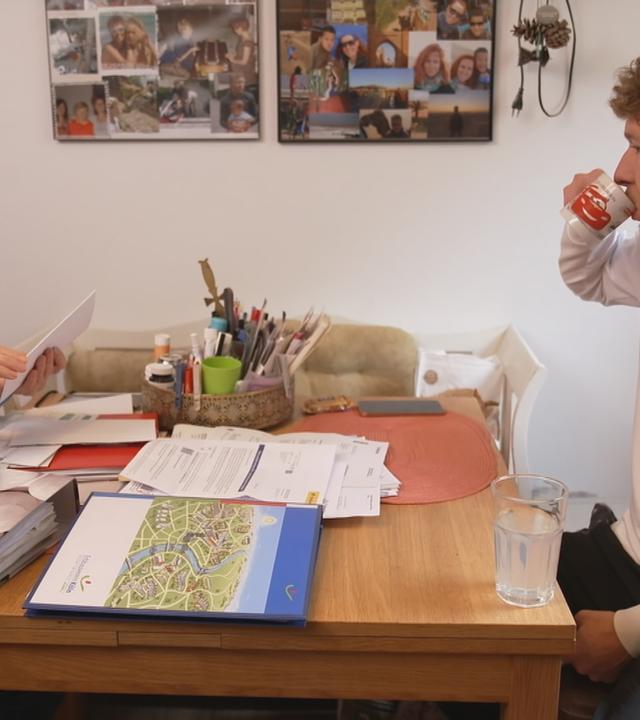 Ein Mann und eine Frau sitzen sich am Tisch gegenüber. Vor ihnen liegen Papierstapel, Ordner und diverse Unterlagen.