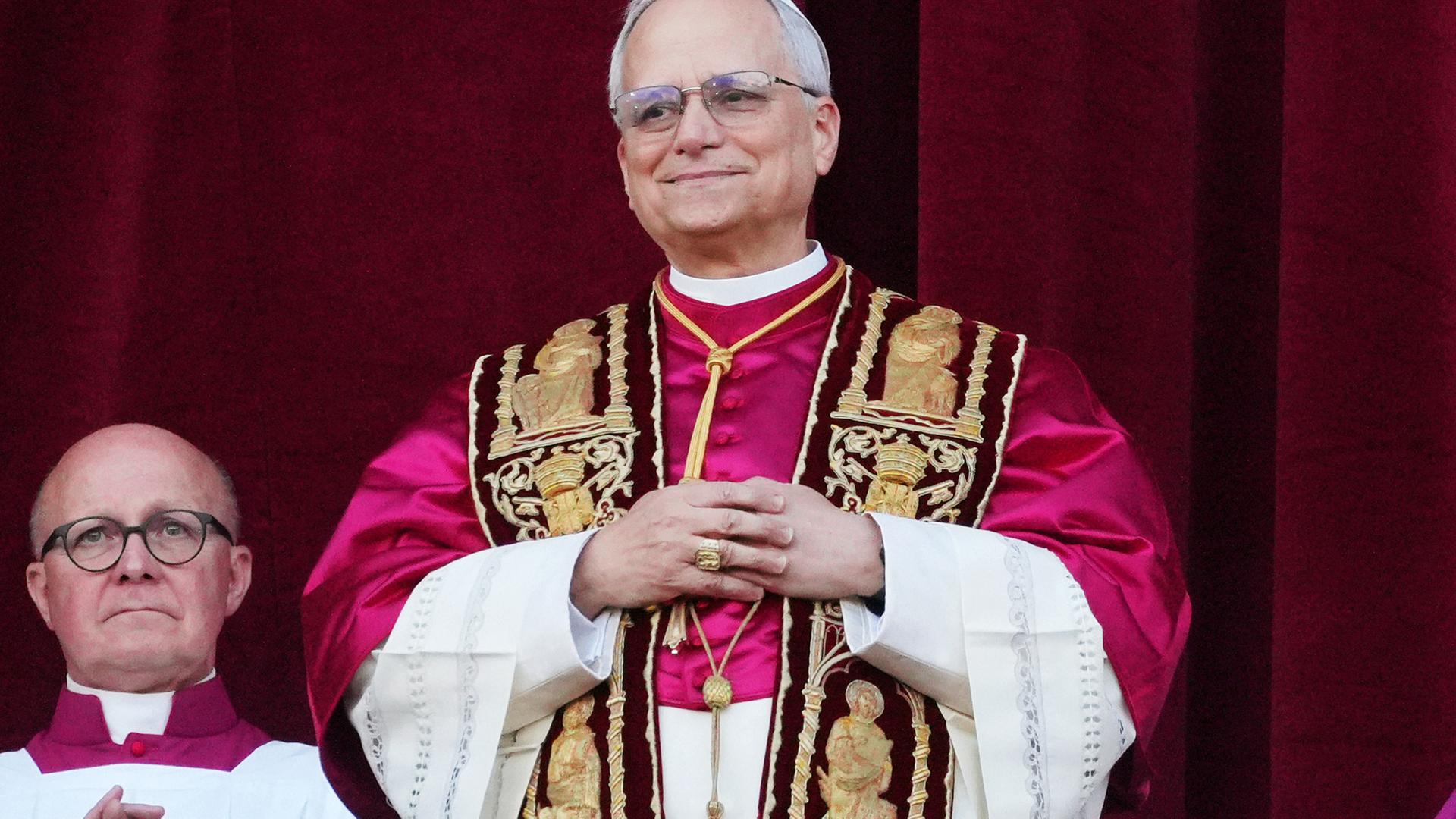 Mann im Papst-Ornat steht lächelnd vor einem purpurnen Vorhang.