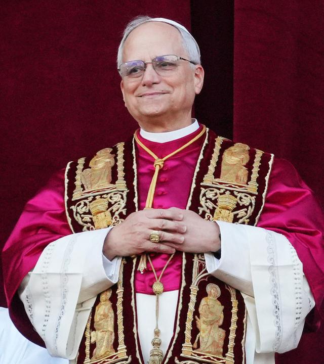 Mann im Papst-Ornat steht lächelnd vor einem purpurnen Vorhang.