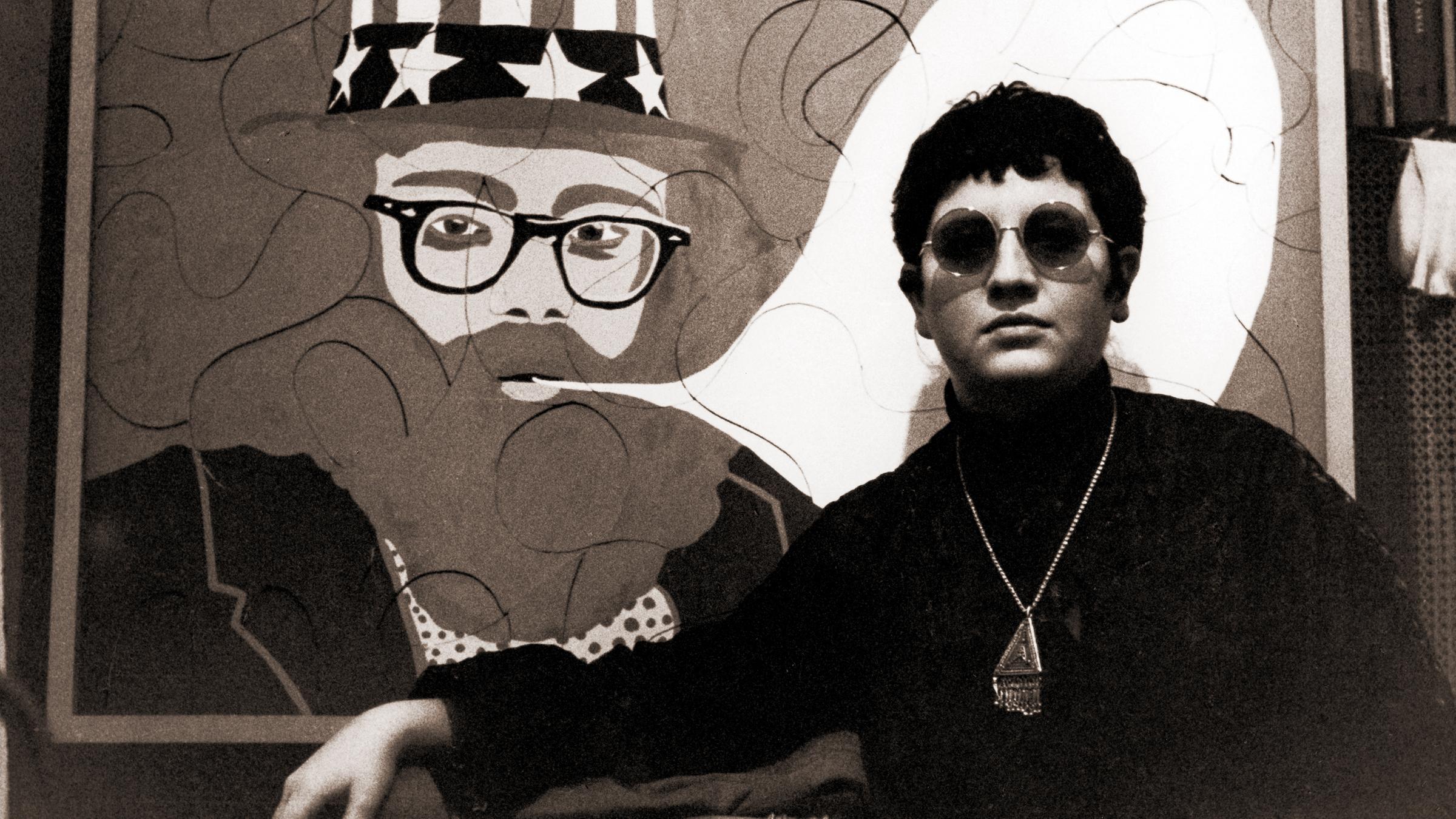 Schwarzweißfoto einer jungen Frau mit kurzen Haaren, einer Sonnenbrille, einem Rollkragenpullover und einer groß perligen Kette um den Hals, die vor einem Bild im Pop Art-Stil steht, das den Dichter Alan Ginsburg mit einer leeren Sprechblase zeigt.