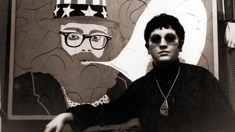 Schwarzweißfoto einer jungen Frau mit kurzen Haaren, einer Sonnenbrille, einem Rollkragenpullover und einer groß perligen Kette um den Hals, die vor einem Bild im Pop Art-Stil steht, das den Dichter Alan Ginsburg mit einer leeren Sprechblase zeigt.