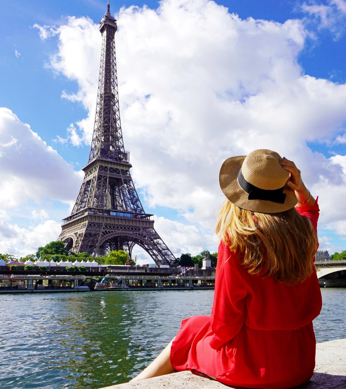 Eine Frau sitzt mit dem Rücken zur Kamera am Ufer der Seine. Sie blickt auf den Eiffelturm.