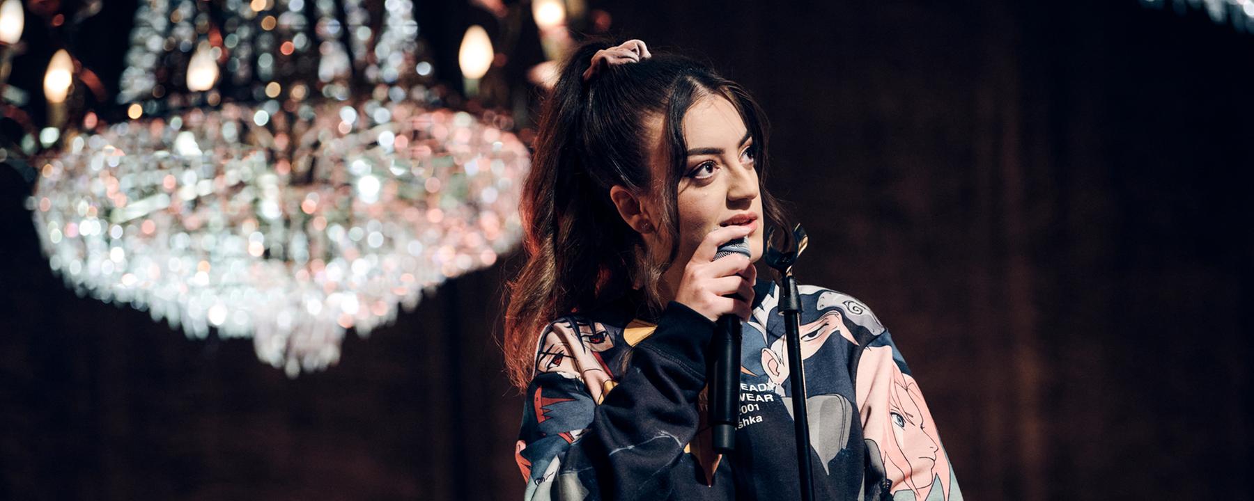 Parshad steht auf einer Bühne, über er ein heller Kronleuchter hängt. Sie hält ein Mikrofon in der Hand. 