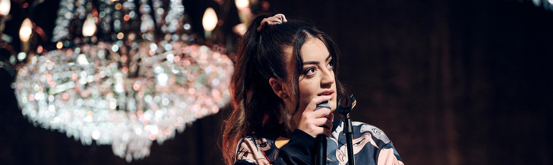 Parshad steht auf einer Bühne, über er ein heller Kronleuchter hängt. Sie hält ein Mikrofon in der Hand. 