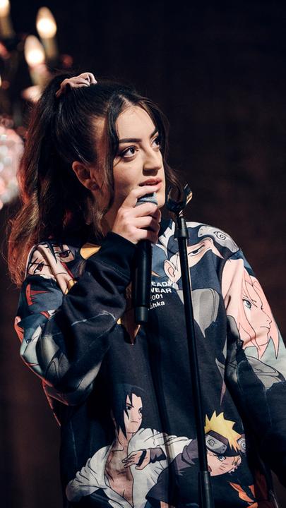 Parshad steht auf einer Bühne, über er ein heller Kronleuchter hängt. Sie hält ein Mikrofon in der Hand. 