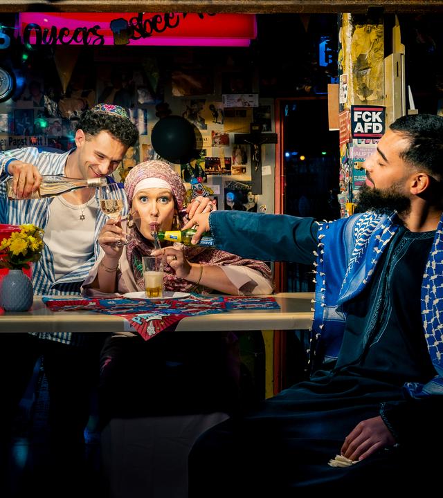 Atheistin Josi sitzt hinter der Bar und trinkt muslimisches Bier aus einem Glas. Vor ihr sitzt Muslim Omar, der ihr das muslimische Bier einschenkt. In der anderen Hand hält Josi ein Weinglas. Dieses füllt ihr Jude Lars auf.