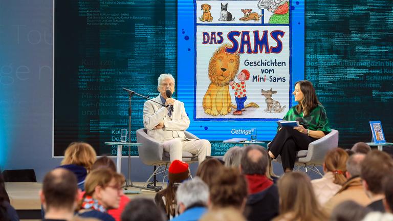 Ein älterer Mann und eine Frau sitzen auf einer Bühne in Sesseln und unterhalten sich vor Publikum. Im Hintergrund ist ein Kinderbuch eingeblendet.