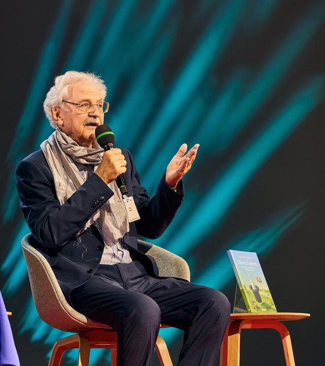 Ein älterer Mann sitzt auf einer Bühne und spricht in ein Mikrofon. Neben ihm sitzt eine Frau mit Mikro in der Hand.