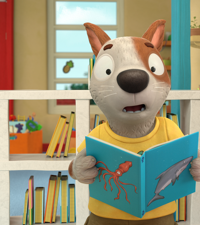 Zac hält ein Buch in den Pfoten und sieht geschockt aus. Auf dem Buch sind ein Krake und ein Hai abgebildet. Im Hintergrund steht ein Regal mit vielen Büchern.