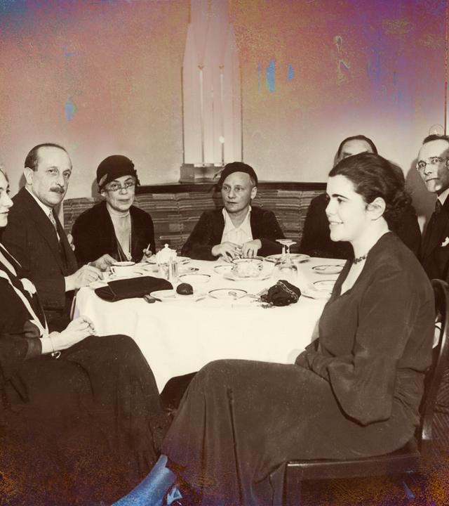 Auf dem sepiafarbenen Foto von 1932 ist eine Gruppe Männer und Frauen an einem runden Tisch zu sehen.