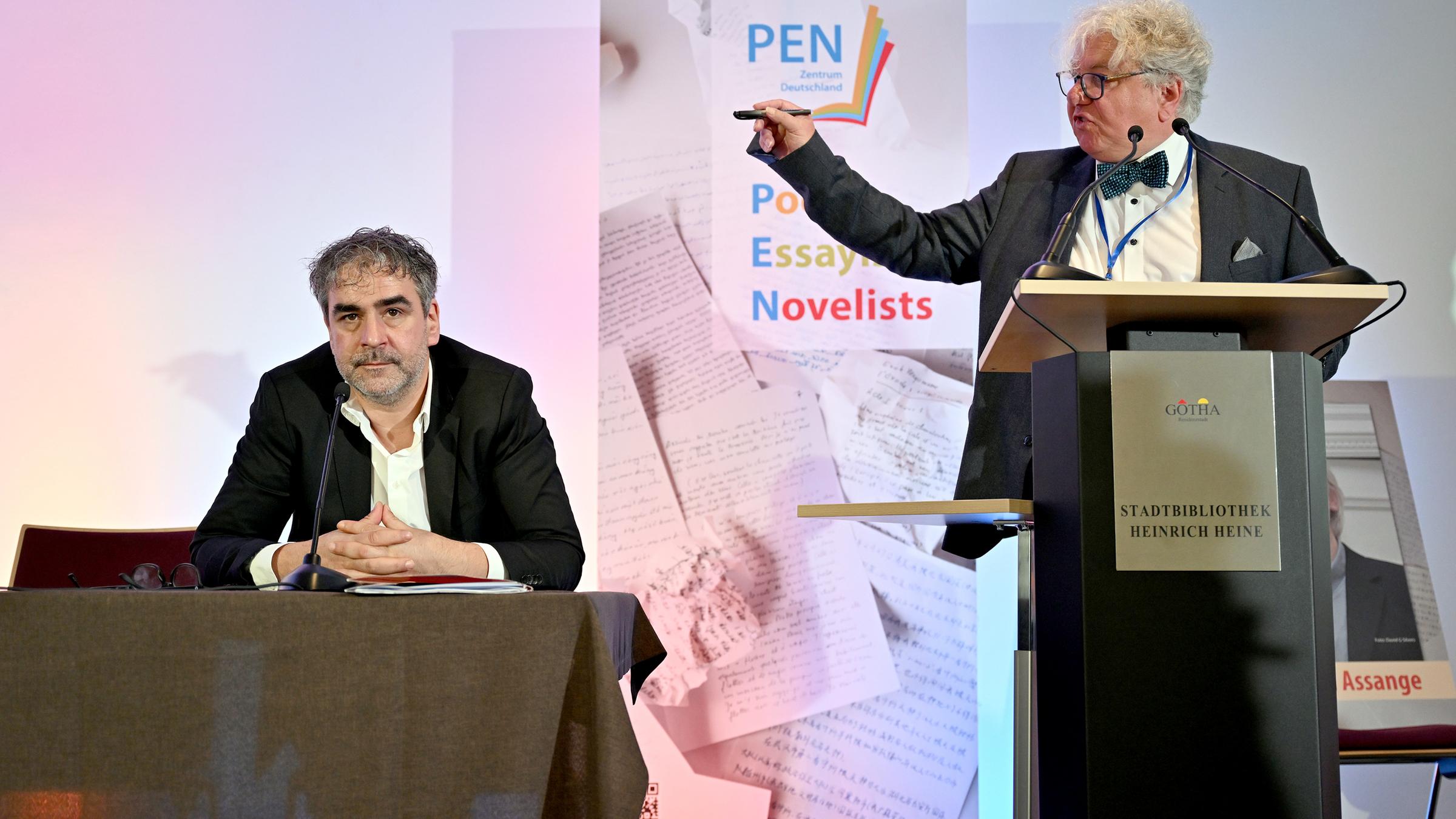 Der PEN--Vorsitzende Deniz Yücel sitzt bei der Pen-Mitgliederversammlung an einem Podest, während PEN-Mitglied Christoph Nix neben ihm an einem Podest wild gestikulierend redet.