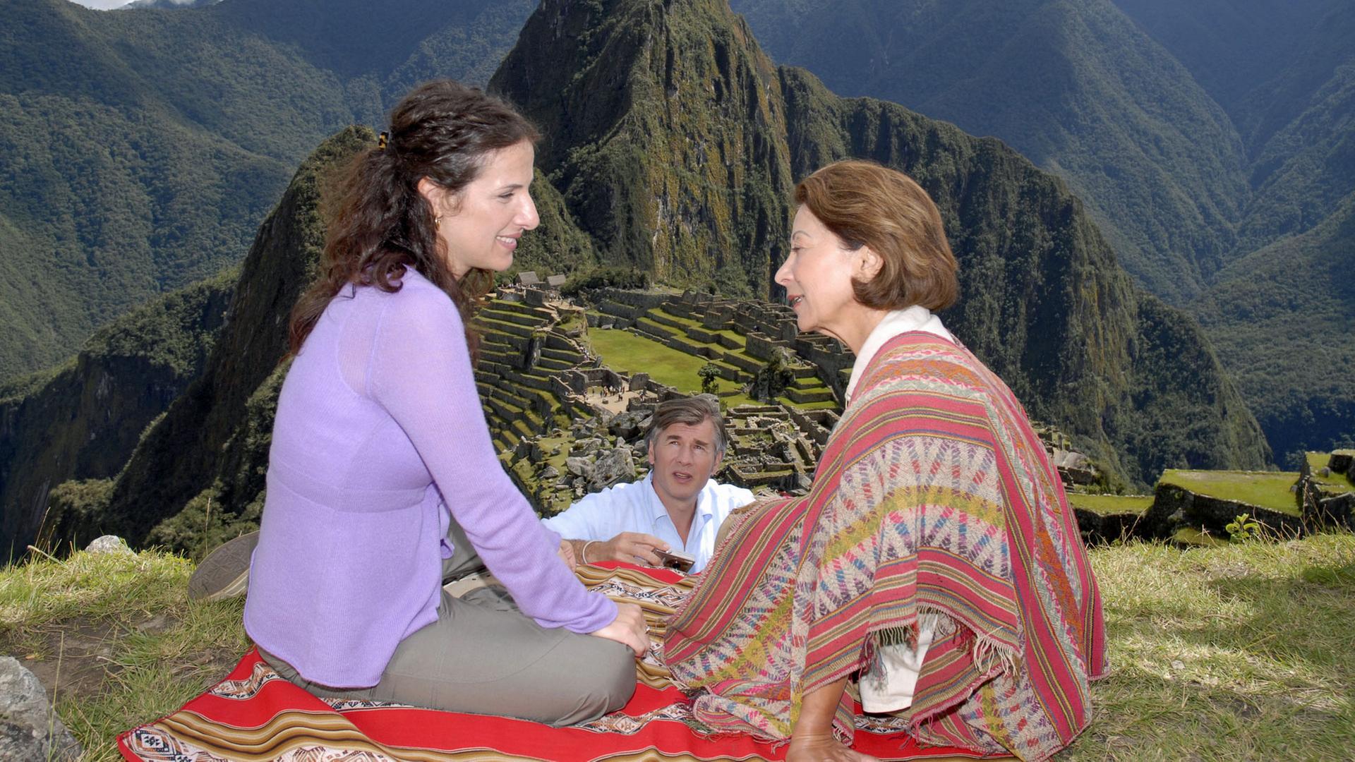 "Das Traumschiff: Peru - Miami": Mara (Clelia Sarto) sitzt mit Consuela (Monica Bielenstein) auf einer bunten Decke auf einer Wiese. Sie unterhalten sich lächelnd. Florian (Daniel Morgenroth) steht etwas abseits, hält eine Kamera in der Hand und beobachtet die beiden. Im Hintergrund sieht man die Inka-Stätte Machu Picchu und die grünbewachsenen Berge Perus.