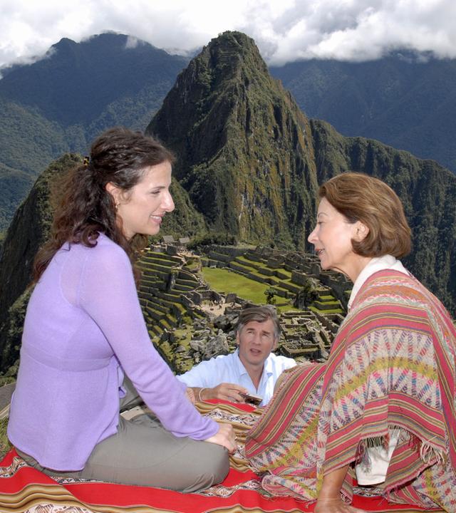 "Das Traumschiff: Peru - Miami": Mara (Clelia Sarto) sitzt mit Consuela (Monica Bielenstein) auf einer bunten Decke auf einer Wiese. Sie unterhalten sich lächelnd. Florian (Daniel Morgenroth) steht etwas abseits, hält eine Kamera in der Hand und beobachtet die beiden. Im Hintergrund sieht man die Inka-Stätte Machu Picchu und die grünbewachsenen Berge Perus.
