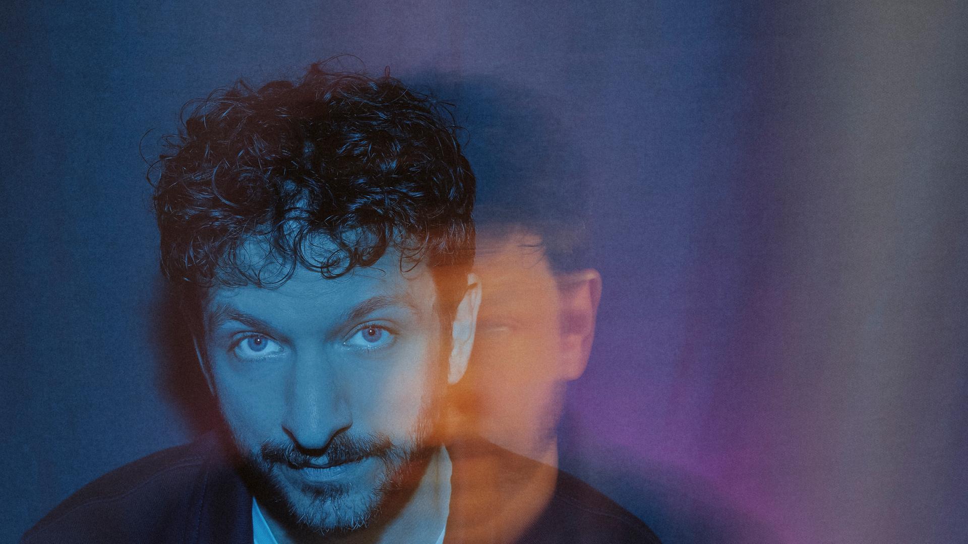 Porträt eines Musikers mit farbigem Lichtspiel im Hintergrund; künstlerisch inszenierte Aufnahme mit weichen Farbverläufen in Blau und Rot.