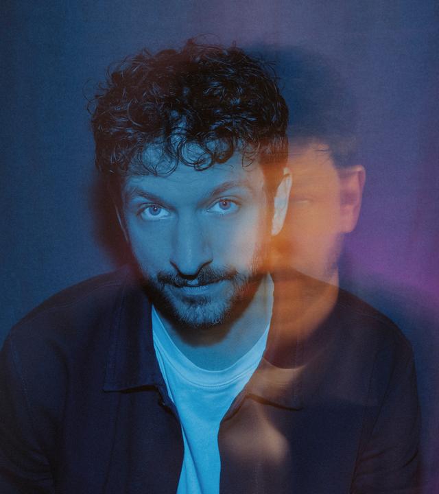 Porträt eines Musikers mit farbigem Lichtspiel im Hintergrund; künstlerisch inszenierte Aufnahme mit weichen Farbverläufen in Blau und Rot.