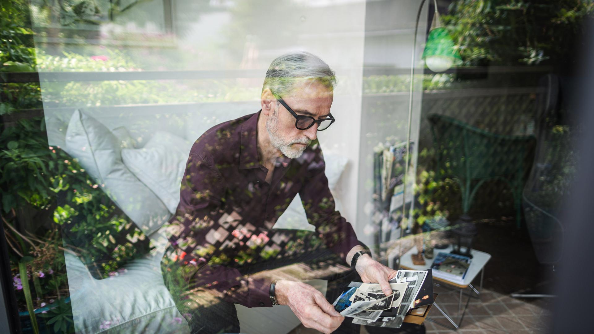 Blick durch eine spiegelnde Scheibe: Ein Mann mit geraumeliertem Haar, Bart und dunkler Brille sitzt auf einem Sofa und betrachtet Fotos.
