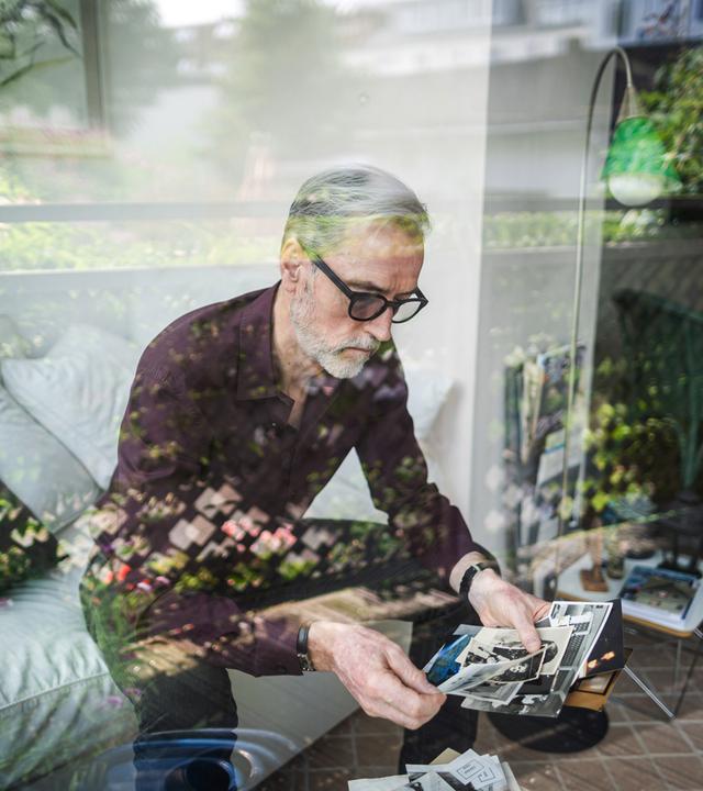 Blick durch eine spiegelnde Scheibe: Ein Mann mit geraumeliertem Haar, Bart und dunkler Brille sitzt auf einem Sofa und betrachtet Fotos.