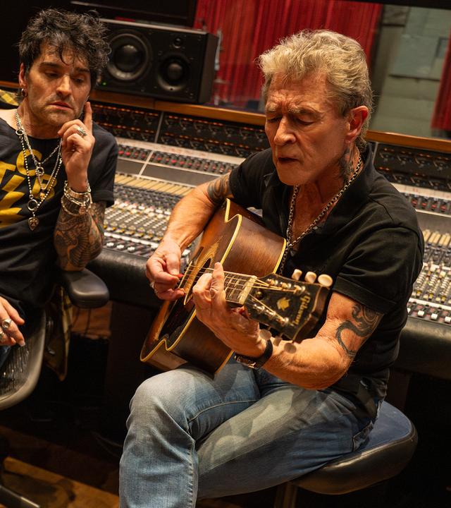 Peter Maffay sitzt auf einem Stuhl, spielt Gitarre und singt. Hinter ihm ist ein großes Mischpult und neben ihm sitzt ein Mann, der Peter Maffay anschaut.
