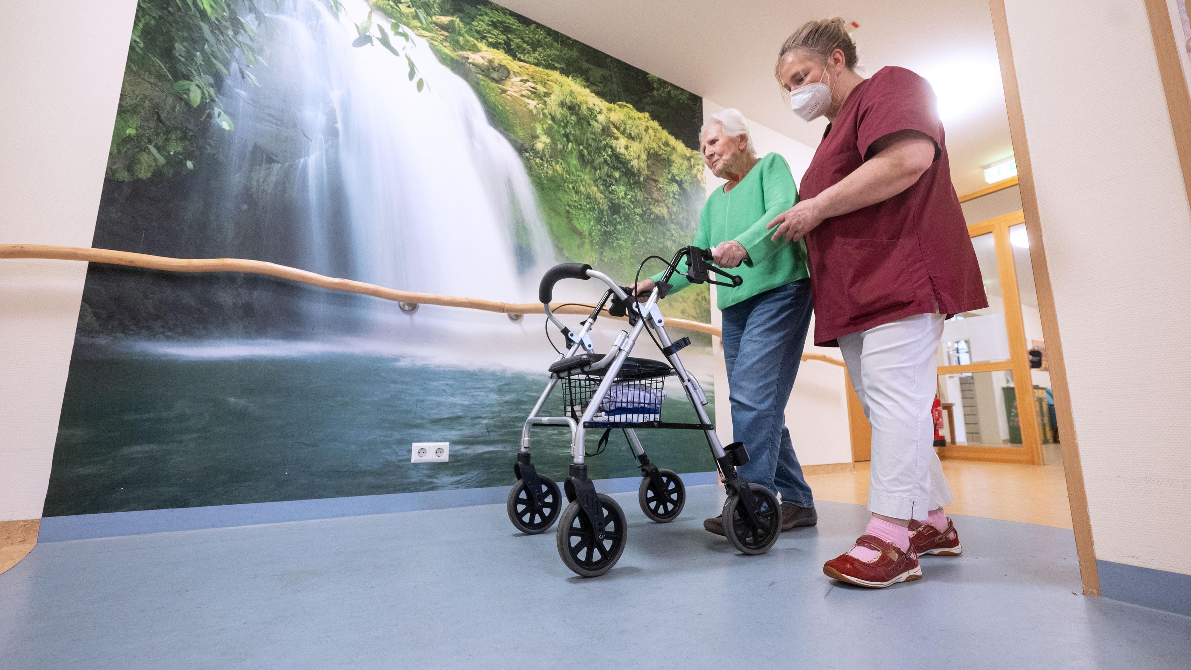 Eine Altenpflegerin hilft älterer Frau im Seniorenheim dabei, mit einem Rollator zu gehen. Im Hintergrund ist ein Bild von einem Wasserfall.
