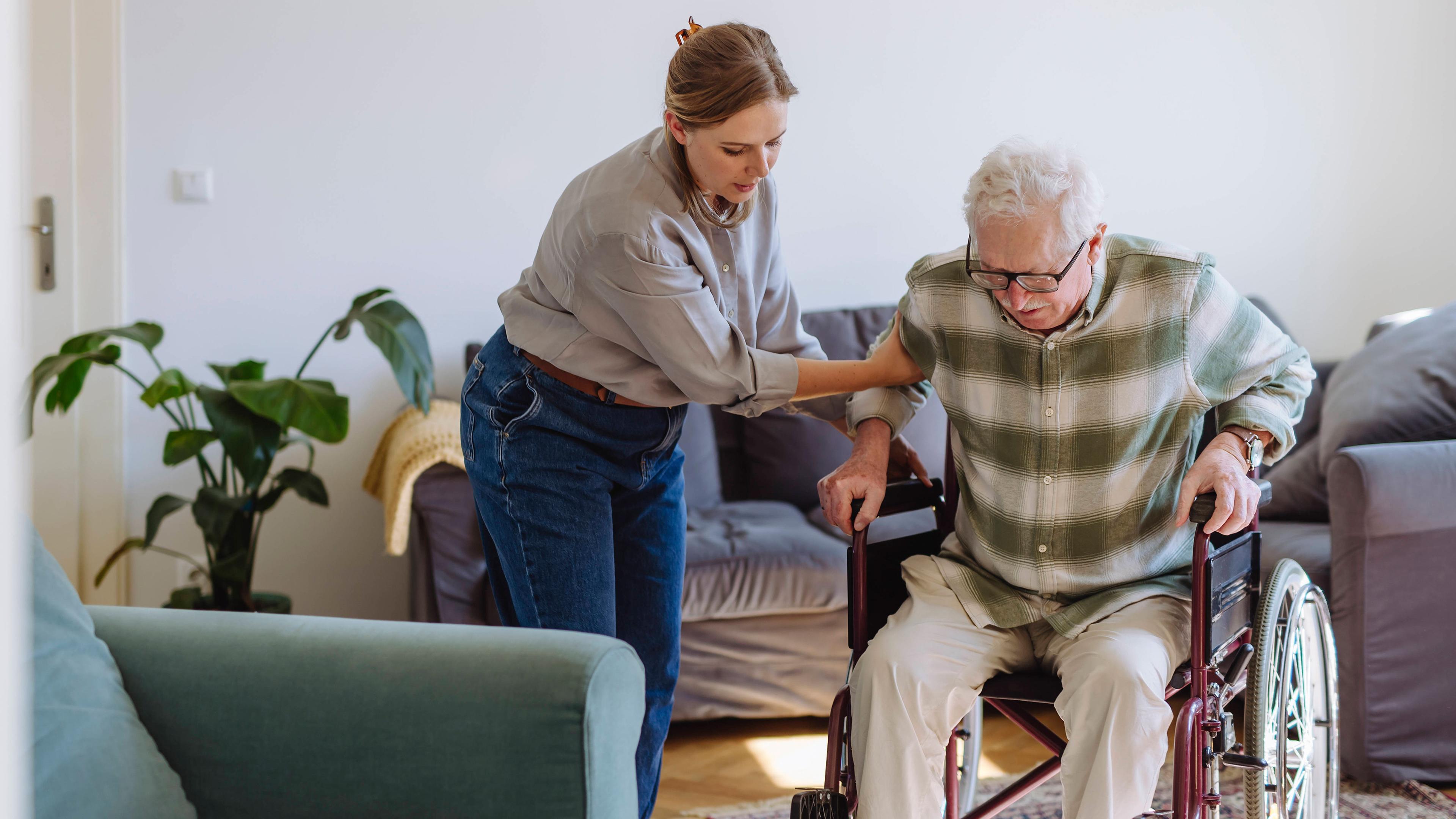 Eine Frau hilft älteren Mann im Rollstuhl sitzend zu Hause