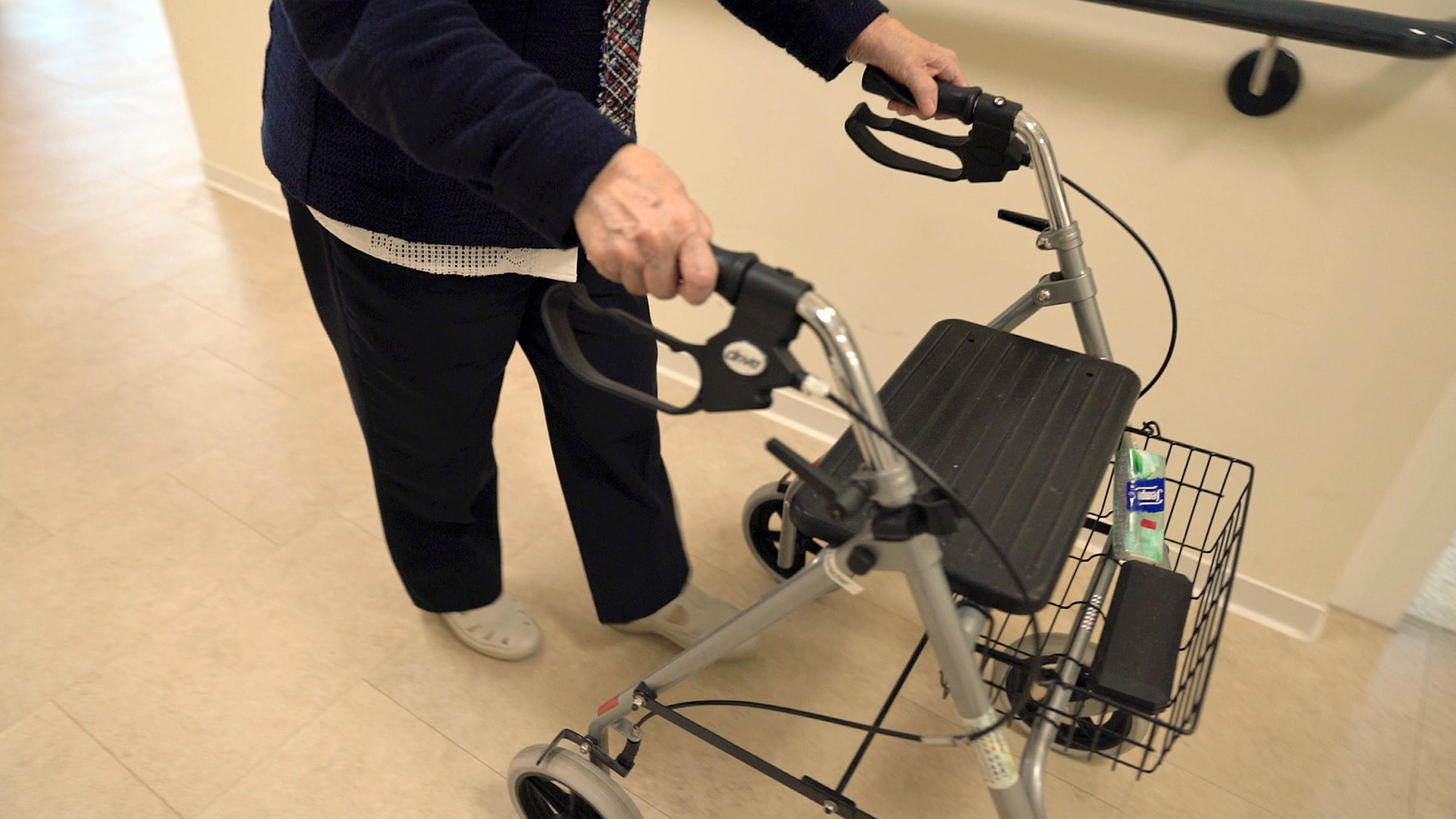 Eine Frau, die man nur von der Schulter abwärts sieht, geht mit Rollator einen Flur entlang.