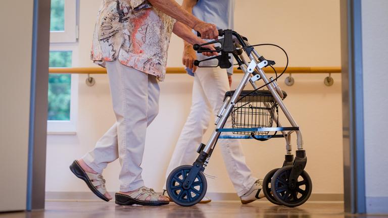 Eine alte Dame schiebt ihren Rollator und wird von einer Pflegekraft begleitet. 