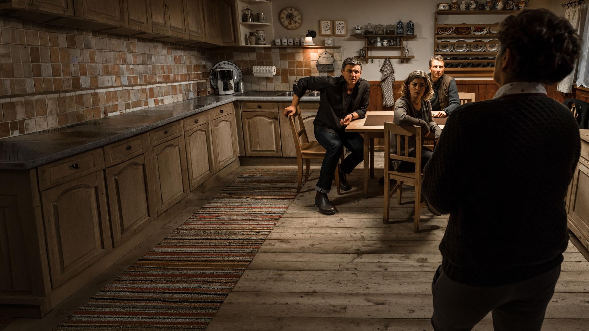 "Der Bergdoktor: Phantomschmerz (1)": Dr. Martin Gruber (Hans Sigl), Lilli Gruber (Ronja Forcher), und Hans Gruber (Heiko Ruprecht) sitzen am Küchentisch. Lisbeth (Monika Baumgartner), nur von hinten sichtbar, steht vor ihnen. Alle schauen sie gespannt an.