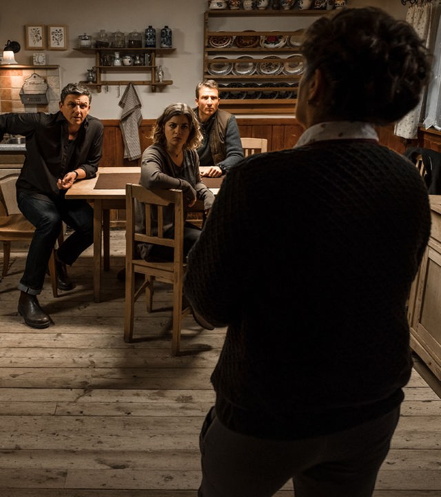 "Der Bergdoktor: Phantomschmerz (1)": Dr. Martin Gruber (Hans Sigl), Lilli Gruber (Ronja Forcher), und Hans Gruber (Heiko Ruprecht) sitzen am Küchentisch. Lisbeth (Monika Baumgartner), nur von hinten sichtbar, steht vor ihnen. Alle schauen sie gespannt an.