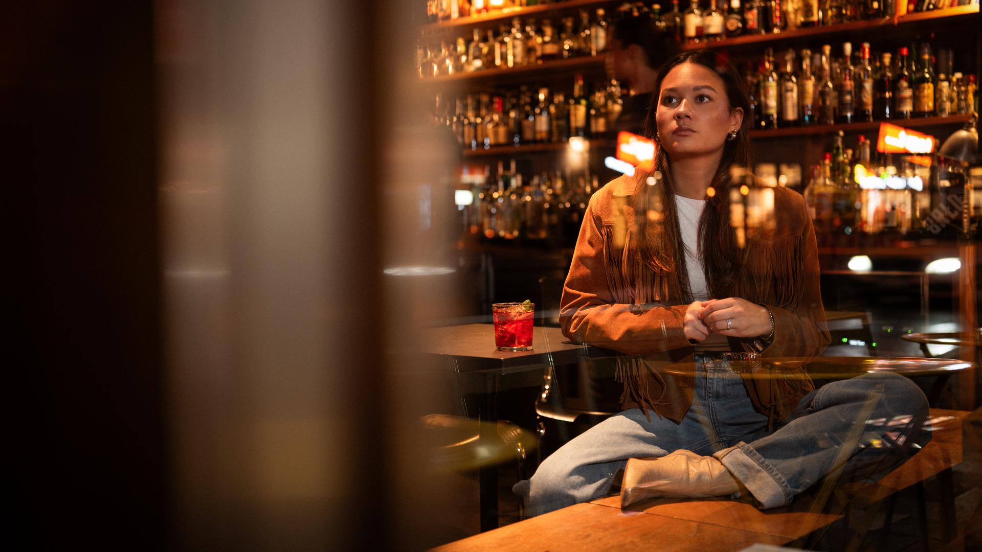 Leonie, eine junge Frau, sitzt in einer Bar auf einer Bank und schaut gedankenverloren aus dem Fenster. Sie stützt sich auf einen Tisch ab, auf dem ein Getränk steht.