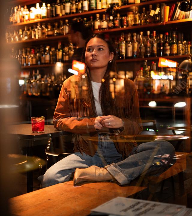 Leonie, eine junge Frau, sitzt in einer Bar auf einer Bank und schaut gedankenverloren aus dem Fenster. Sie stützt sich auf einen Tisch ab, auf dem ein Getränk steht.