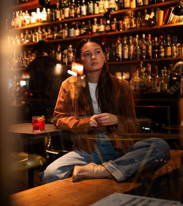 Leonie, eine junge Frau, sitzt in einer Bar auf einer Bank und schaut gedankenverloren aus dem Fenster. Sie stützt sich auf einem Tisch ab, auf dem ein Getränk steht.