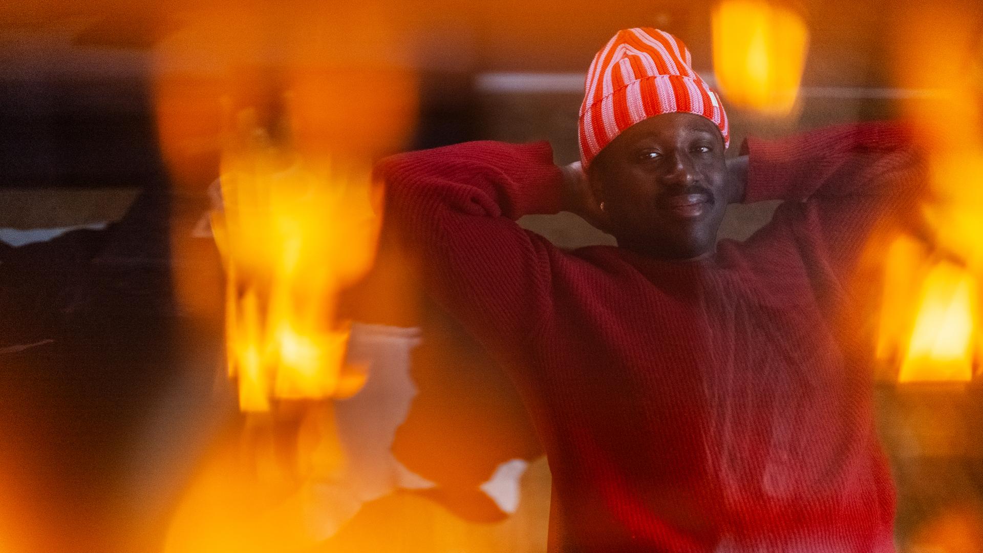 Jonny sitzt auf einem Stuhl und hat die Arme lässig hinter seinem Kopf, um diesen zu stützen. Er lächelt in die Kamera. Das Bild wurde durch eine Lampe durchfotografiert, wodurch Lichteffekte auf dem Bild zu sehen sind. 