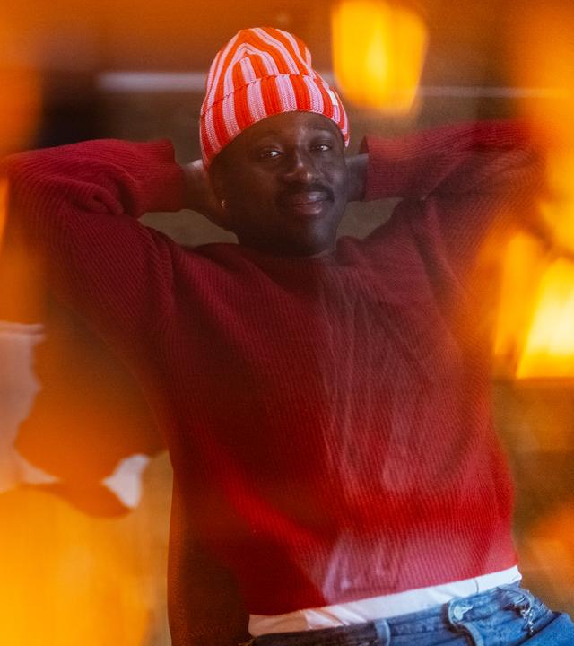 Jonny sitzt auf einem Stuhl und hat die Arme lässig hinter seinem Kopf, um diesen zu stützen. Er lächelt in die Kamera. Das Bild wurde durch eine Lampe durchfotografiert, wodurch Lichteffekte auf dem Bild zu sehen sind. 