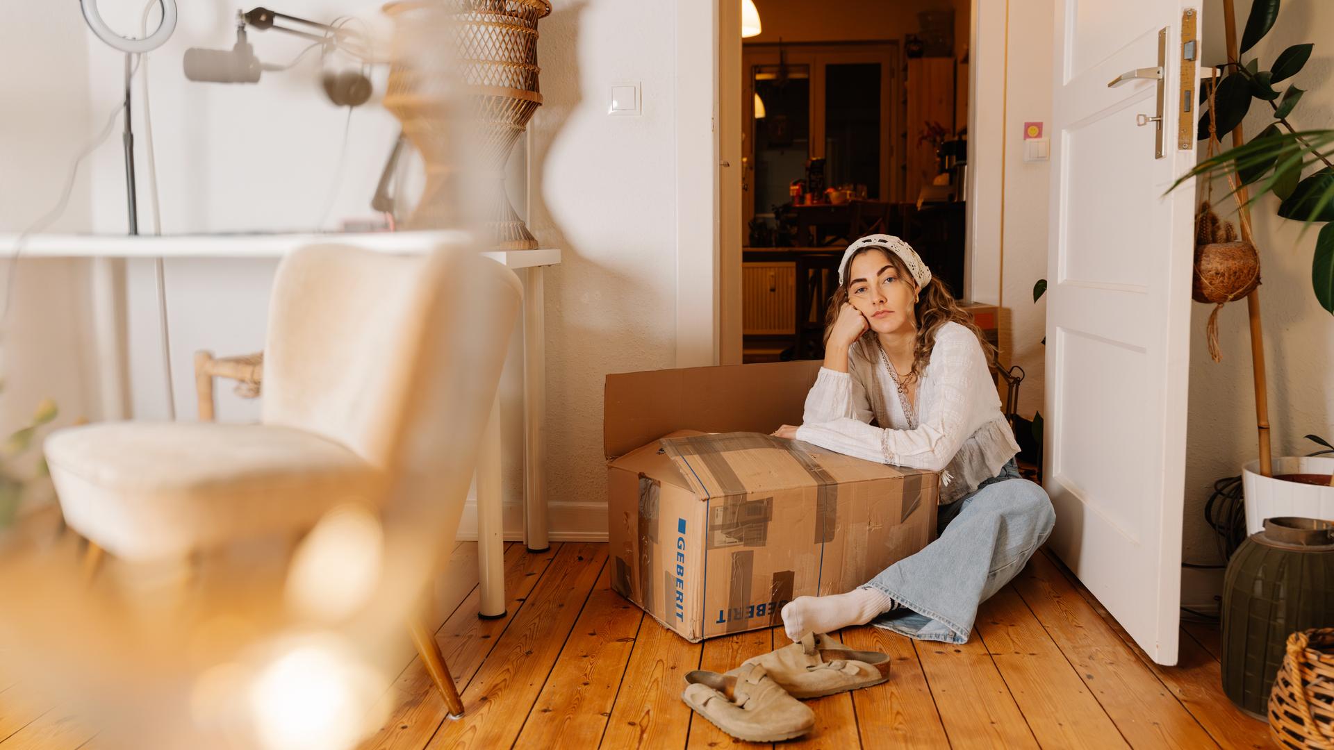 Lisa, eine junge Frau, sitzt auf dem Boden einer Wohnung. Sie stützt sich auf einen Umzugskarton ab und schaut lustlos in die Kamera.