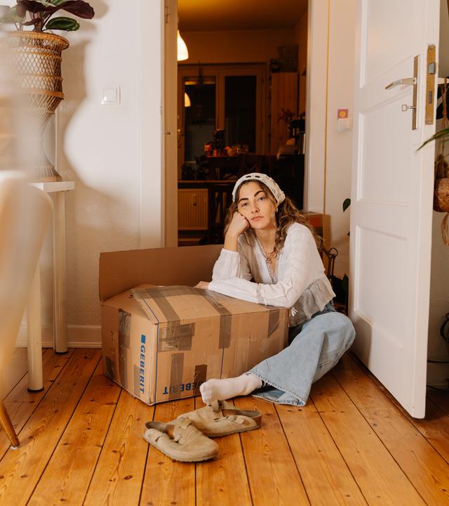 Lisa, eine junge Frau, sitzt auf dem Boden einer Wohnung. Sie stützt sich auf einen Umzugskarton ab und schaut lustlos in die Kamera.