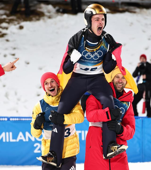 Philipp Raimund (GER) jubelt mit seinen Teamkollegen Felix Hoffmann (GER) und Pius Paschke (GER) nach seinem Sieg auf der Normalschanze, Durchgang 2, Olympische Winterspiele 2026.