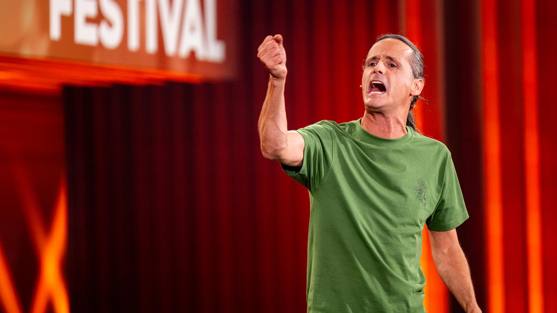 Philipp Weber steht auf der rechten Seite des Bildes. Mit seiner rechten Hand deutet er in Richtung des linken Bildrandes. Er trägt ein grünes T-Shirt. Der Hintergrund ist rot