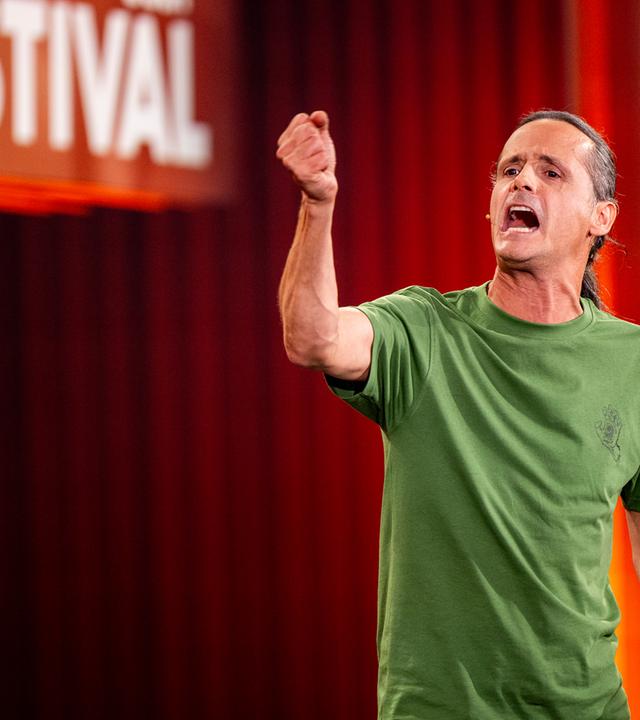 Philipp Weber steht auf der rechten Seite des Bildes. Mit seiner rechten Hand deutet er in Richtung des linken Bildrandes. Er trägt ein grünes T-Shirt. Der Hintergrund ist rot