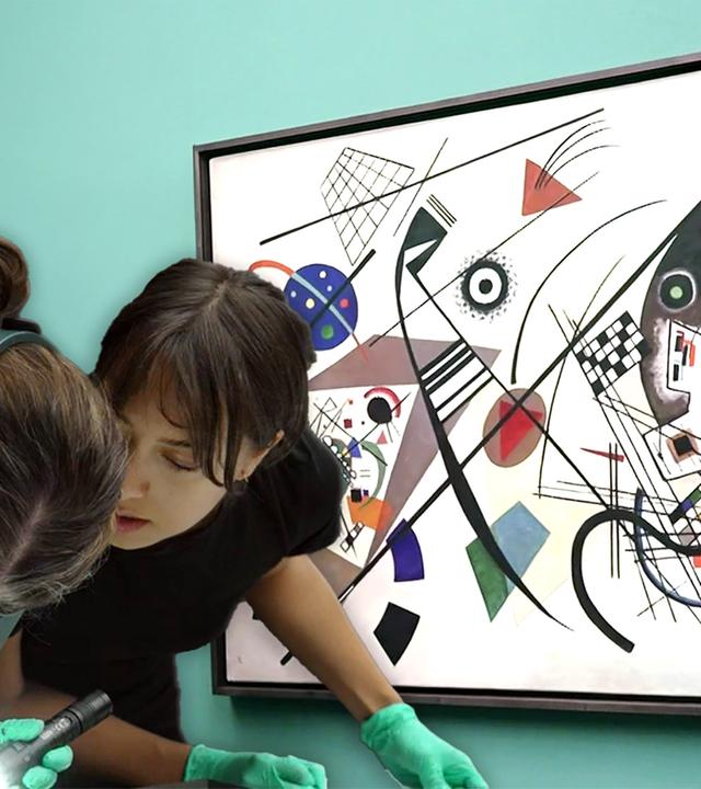 Zwei Frauen beugen sich neben einem an der Wand hängenden Bild von Miró auf ein vor ihnen liegendes Werk.