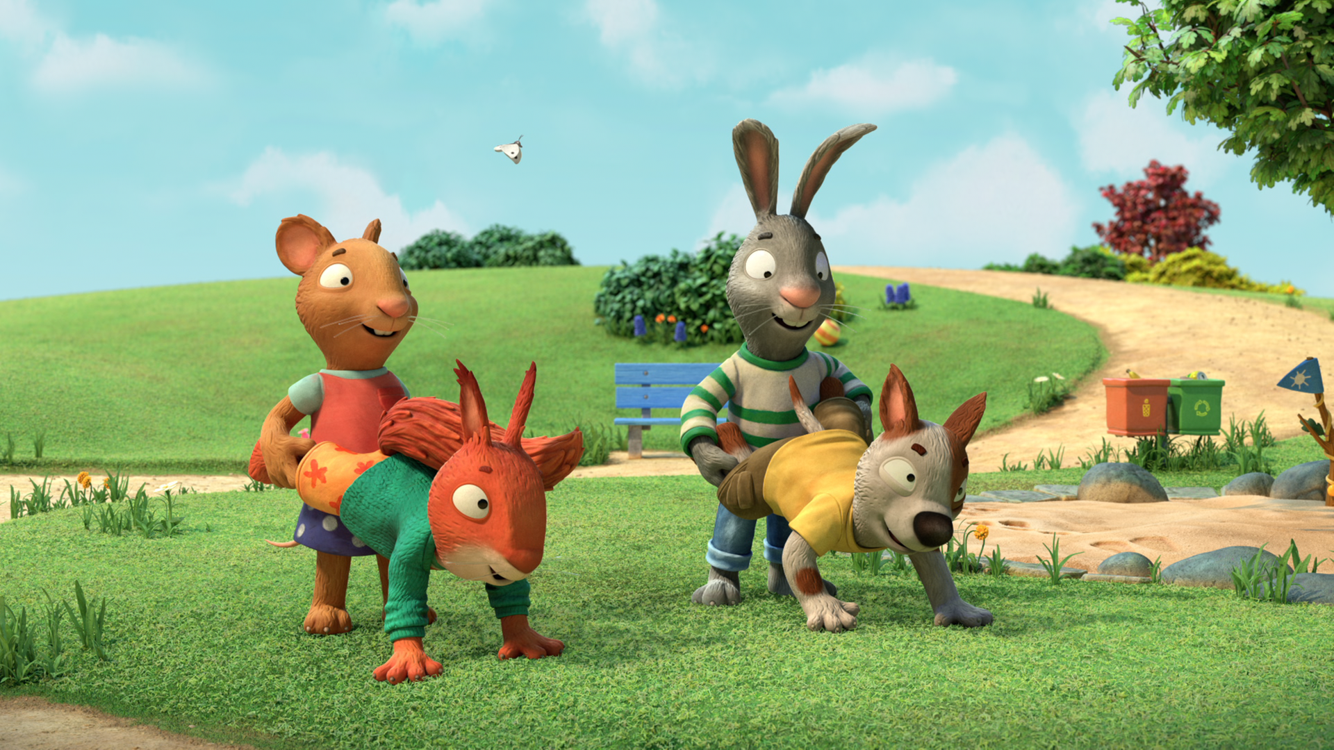 Posy, die Maus und Pip, das Kaninchen stehen nebeneinander. Vor Posy in Schubkarrenposition befindet sich Jamila, das Eichhörnchen und vor Pip, ebenfalls in Schubkarrenposition, befindet sich Zac, der Hund. Sie schauen alle lächelnd und erwartungsvoll. Im Hintergrund sind Sträucher, Blumen, eine blaue Parkbank, zwei Mülleimer und ein Sandkasten zu sehen.