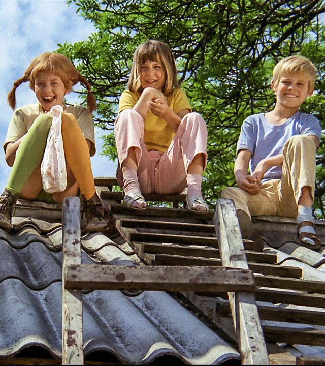 Pippi Langstrumpf sitzt mit Annika und Tommy auf dem Dach. Sie lachen alle drei. Eine Leiter steht angeleihnt davor. 