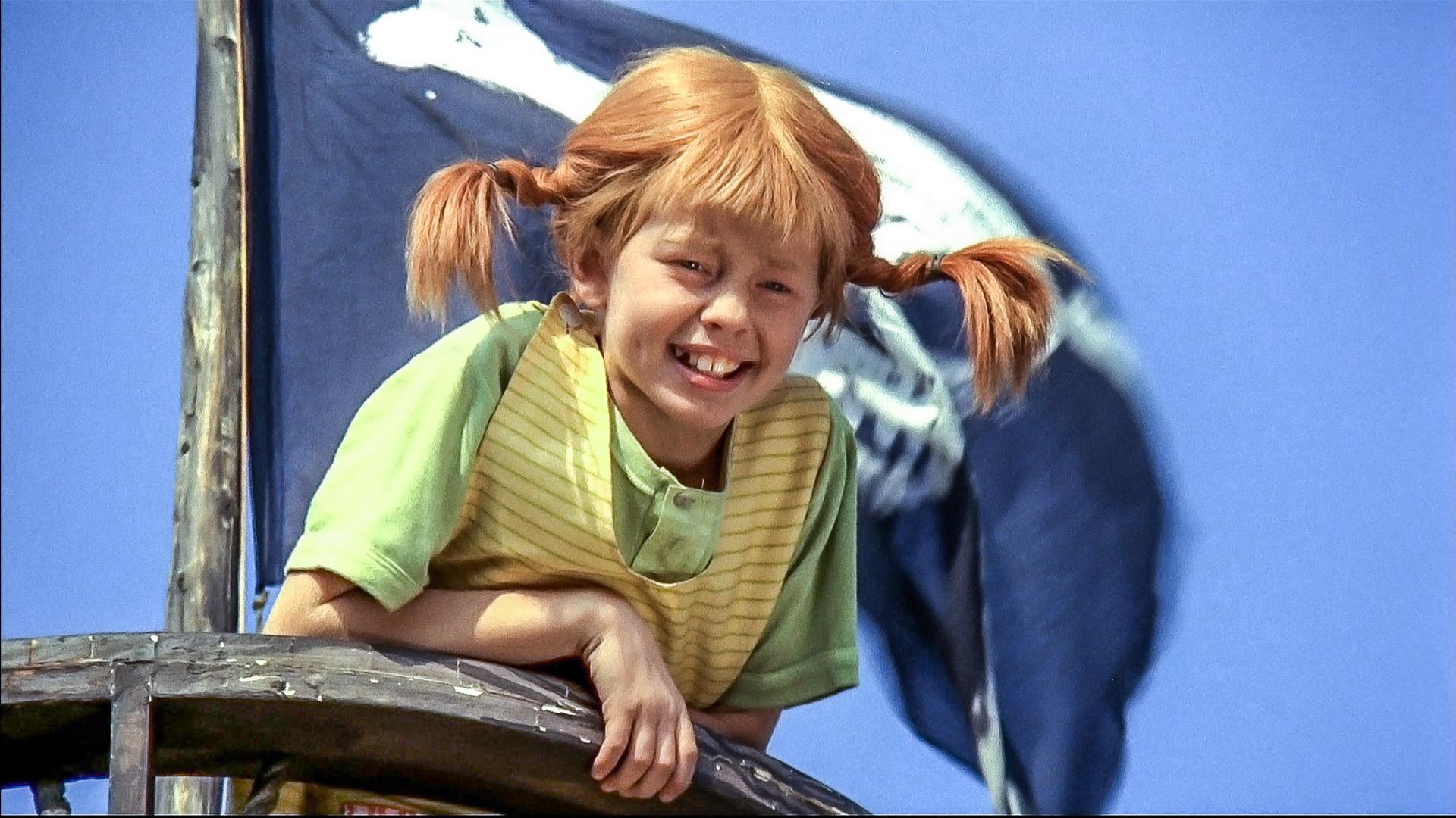 Pippi Langstrumpf ist hoch oben im sogenannten Krähennest, dem Mastkorb eines hölzernen Segelschiffs. Hinter ihr weht die Piratenflagge am Mast.