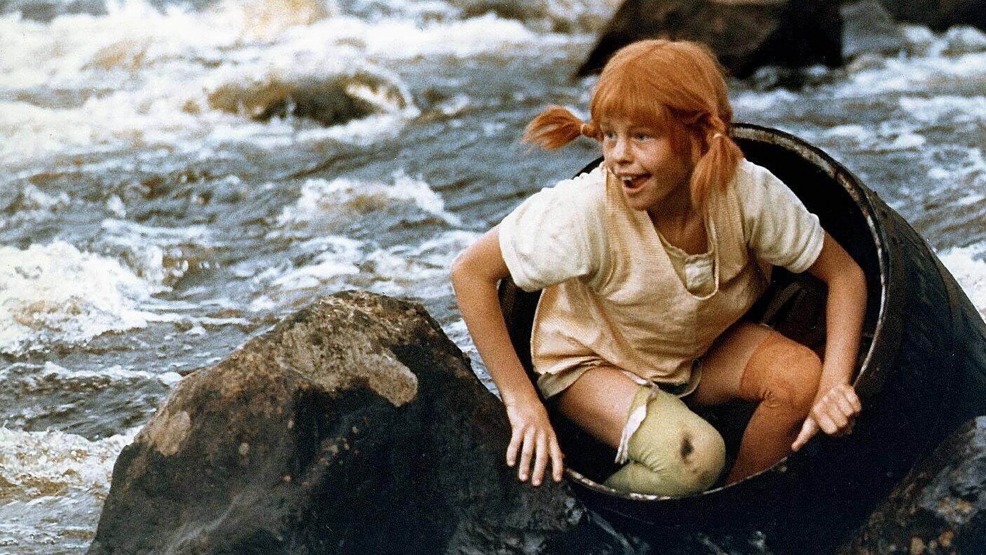 Pippi Langstrumpf sitzt in einem Fass, das in einem Fluss schwimmt. Es ragen Felsen aus dem Wasser. 