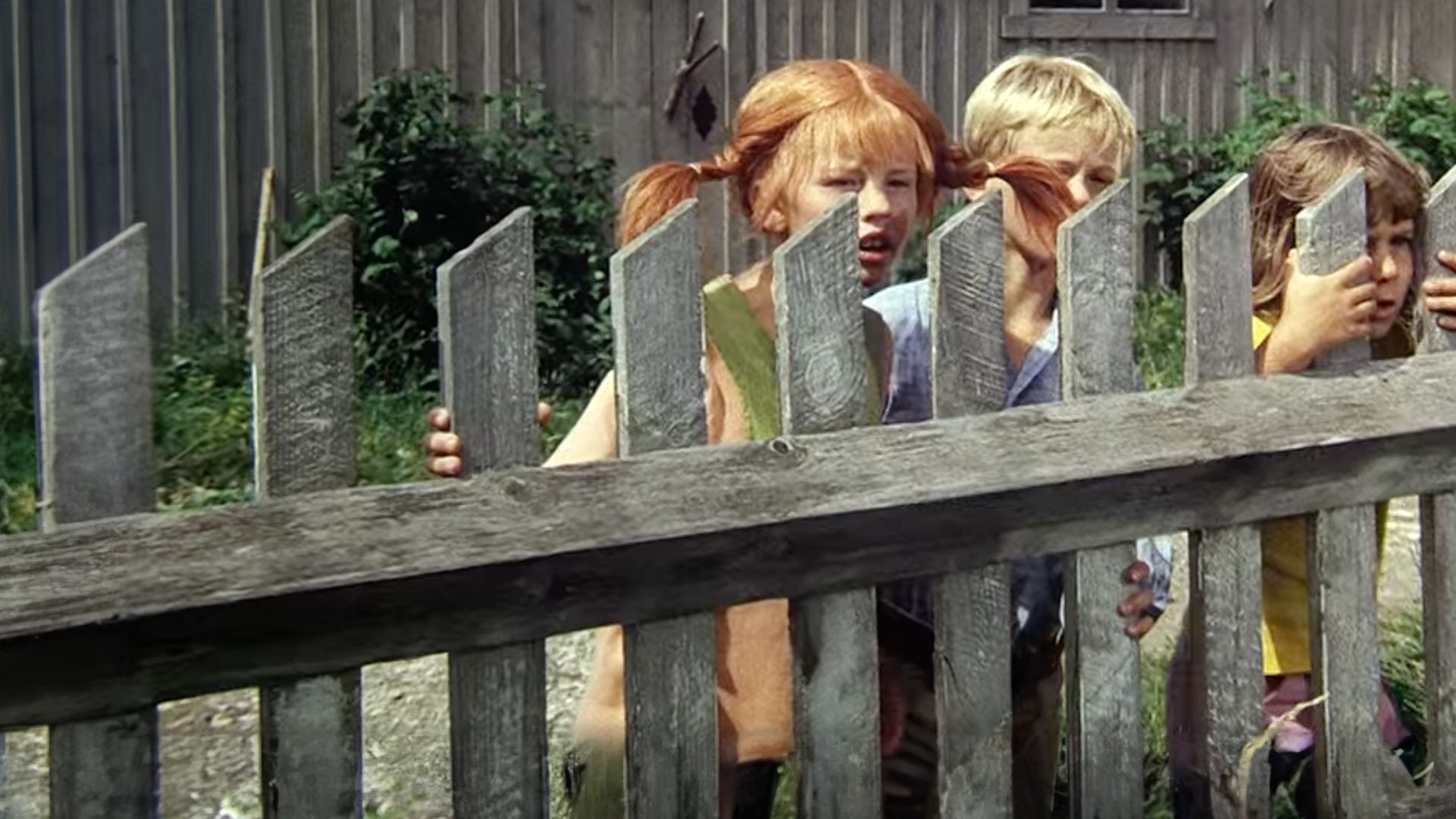 Pippi Langstrumpf steht versteckt mit Tommy und Annika hinter einem Bretterzaun.