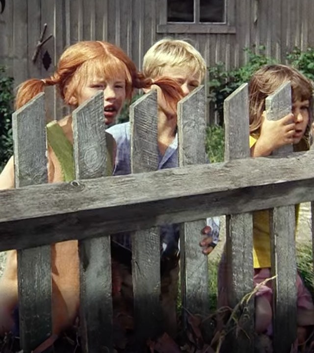 Pippi Langstrumpf steht versteckt mit Tommy und Annika hinter einem Bretterzaun.