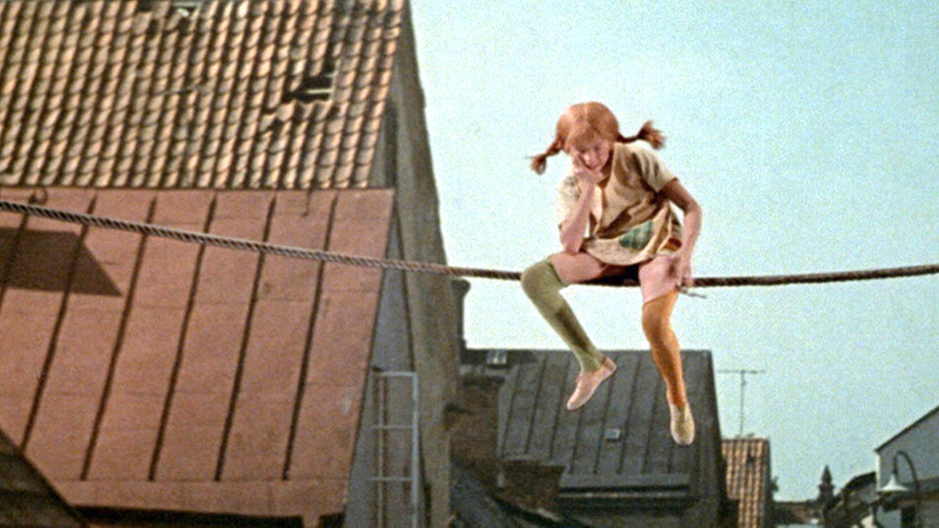 Pippi sitzt auf einem Seil, welches zwischen den Häuserdächern gespannt ist, in der Luft.