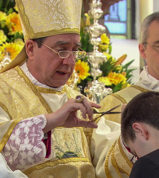 Das Bild zeigt eine Zeremonie in einem religiösen Kontext. Im Vordergrund ist ein junger Mann zu sehen, der sich leicht nach vorne beugt. Ein Bischof, gekleidet in eine prunkvolle, goldene liturgische Kleidung mit einem Mitra auf dem Kopf, schneidet mit einer Schere eine kleine Haarsträhne vom Kopf des jungen Mannes ab. Der Bischof hält die Schere in seiner rechten Hand, während er mit der linken Hand eine Handbewegung macht.  Im Hintergrund sind dekorative Blumenarrangements, überwiegend gelb, zu sehen. Neben dem Bischof stehen zwei andere Männer in ähnlicher, aber weniger aufwendiger liturgischer Kleidung, die aufmerksam auf die Zeremonie blicken. Die Atmosphäre wirkt feierlich und rituell.